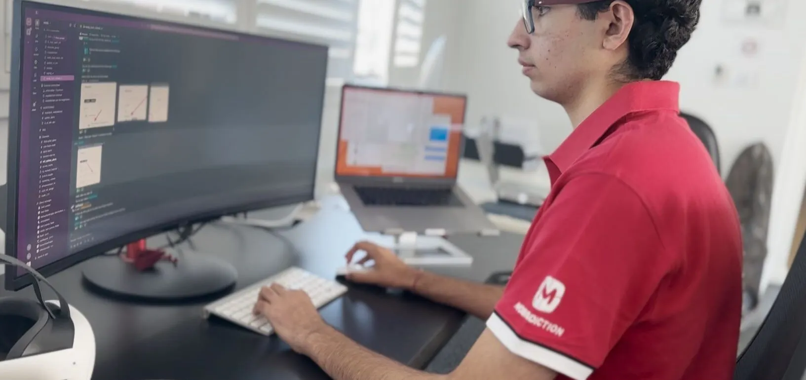 Person in a red polo shirt and glasses sits at a desk, typing while a curved monitor displays code on the screen.