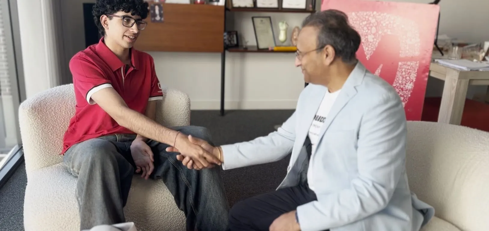 Two men sit on a curved white couch and shake hands in a casual office lounge, smiling at each other after a meeting.